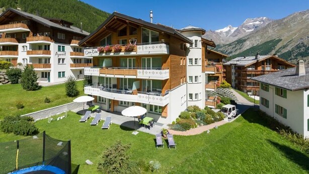 Een hotel met meerdere verdiepingen, balkons, terraszitplaatsen en een bergachtige achtergrond.