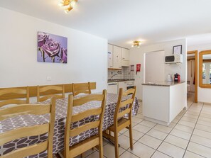 Ein Essbereich mit einem langen Tisch und Stühlen, eine Küche im Hintergrund und ein Gemälde mit violetten Blumen an der Wand.