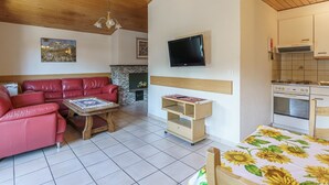 Een woonkamer met een rode leren bank, een houten salontafel, een aan de muur bevestigde televisie, en een keukenhoek met een fornuis en oven.