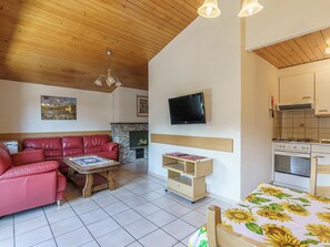 Ein Wohnzimmer mit einem roten Ledersofa, einem Holztisch, einem an der Wand montierten Fernseher und einer Küchenzeile mit Herd und Backofen.