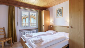 Een kamer met twee bedden, een houten stoel, een raam met gordijnen, een radiator en een schilderij aan de muur.