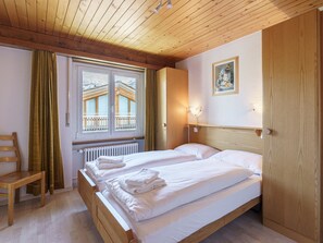 Ein Zimmer mit zwei Betten, einem Holzstuhl, einem Fenster mit Vorhängen, einem Heizkörper und einem Bild an der Wand.
