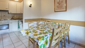 Een eethoek met een tafel voor vier personen, houten stoelen, een keuken op de achtergrond en een ingelijste foto aan de muur.