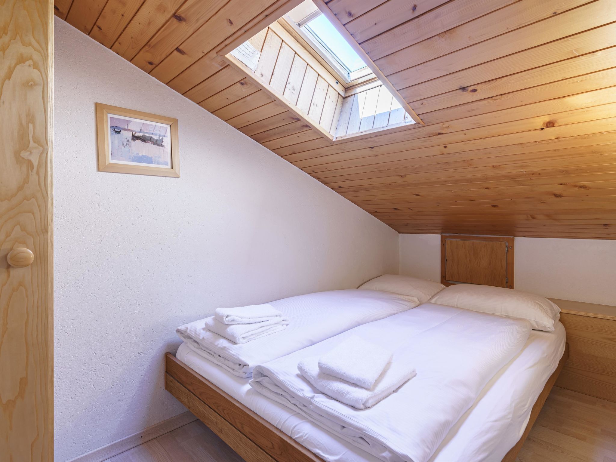 A bedroom with a wooden ceiling, a skylight, a bed with white linens, and a framed picture on the wall.