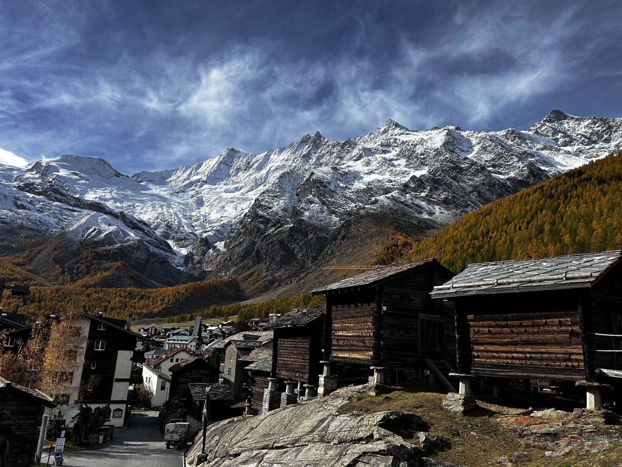 A mountain village with traditional wooden buildings, a clear road, and snow-capped mountains in the background.