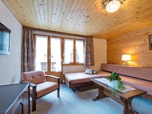 Een knus interieur met houten plafond, een bank, een stoel, een tafeltje met een plant en een raam met gordijnen.