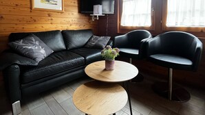 En hyggelig stue med en sort lædersofa, et rundt træbord og to sorte læderlænestole. Der er et fladskærms-tv monteret på væggen, og et vindue med gardiner.