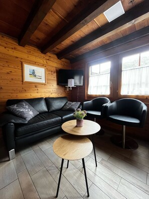 A cozy living room with a black leather sofa, a round wooden coffee table, and two black leather armchairs. A flat-screen TV is mounted on the wall, and there is a window with curtains. - Chalet Heimeli Appartement/Fewo 6-Bettwohnung by Interhome (Saas-Fee)