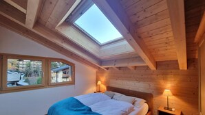 Un dormitorio con tragaluces, paredes de madera, una cama con manta azul, dos lámparas de noche y una ventana con vistas a edificios.