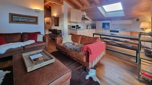Una sala de estar acogedora con un sofá de cuero marrón, una mesa de centro de madera y un tapiz de cuero de vaca. Se ve una cocina al fondo, con armarios blancos y un tragaluz.