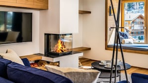 A modern living room with a fireplace, a television, and a window with a view of a mountain lodge. - Riverside 24 Appartement/Fewo Appartement/Fewo by Interhome (Saas-Fee)
