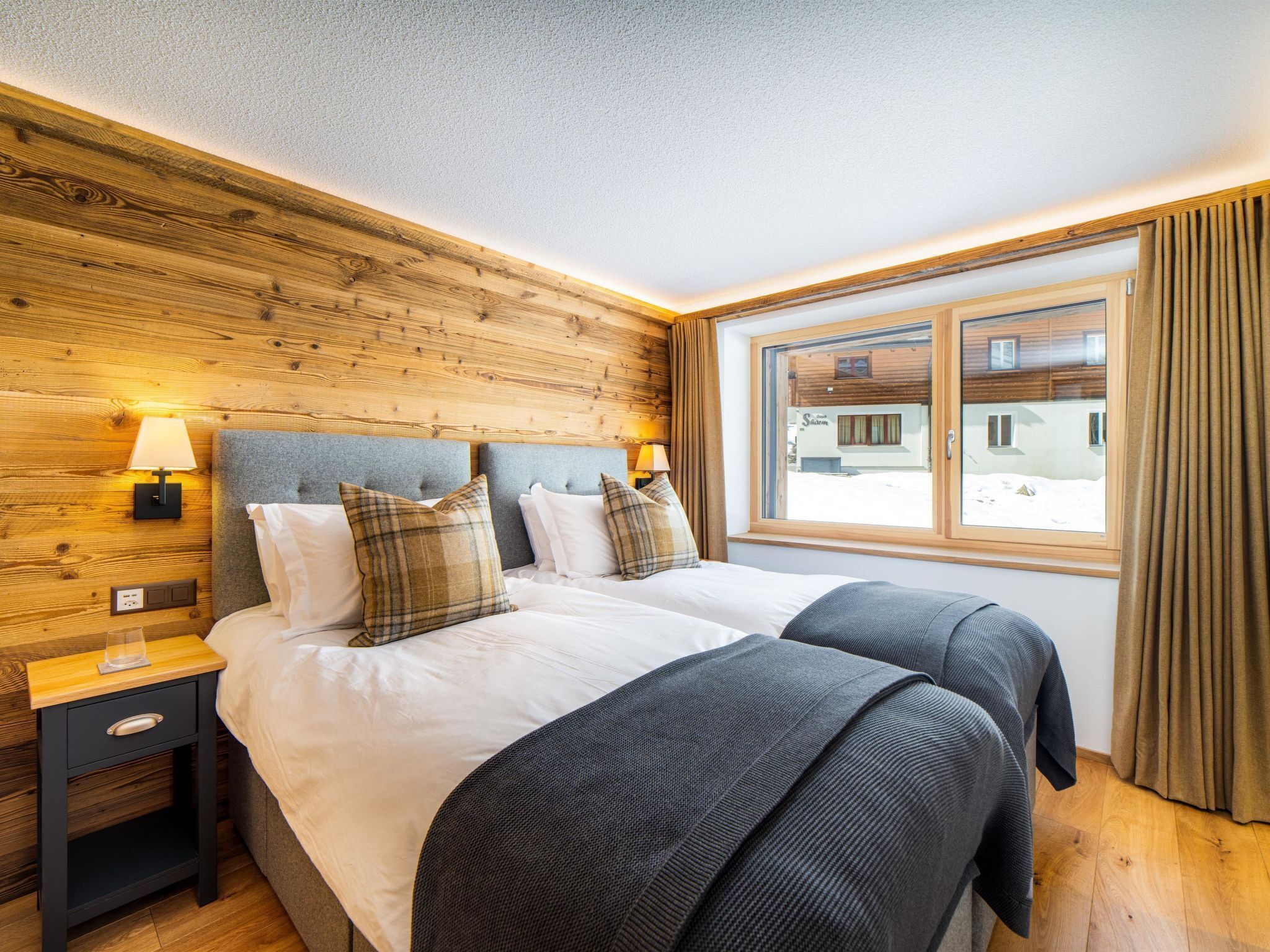 Een tweepersoonsbed met twee kussens, een nachtkastje met een lamp, een houten hoofdbord en een raam met gordijnen.