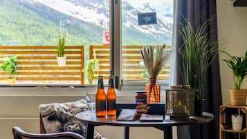 Una habitación acogedora con vistas a montañas nevadas, un balcón de madera con plantas en macetas y una mesa con botellas y una vela.