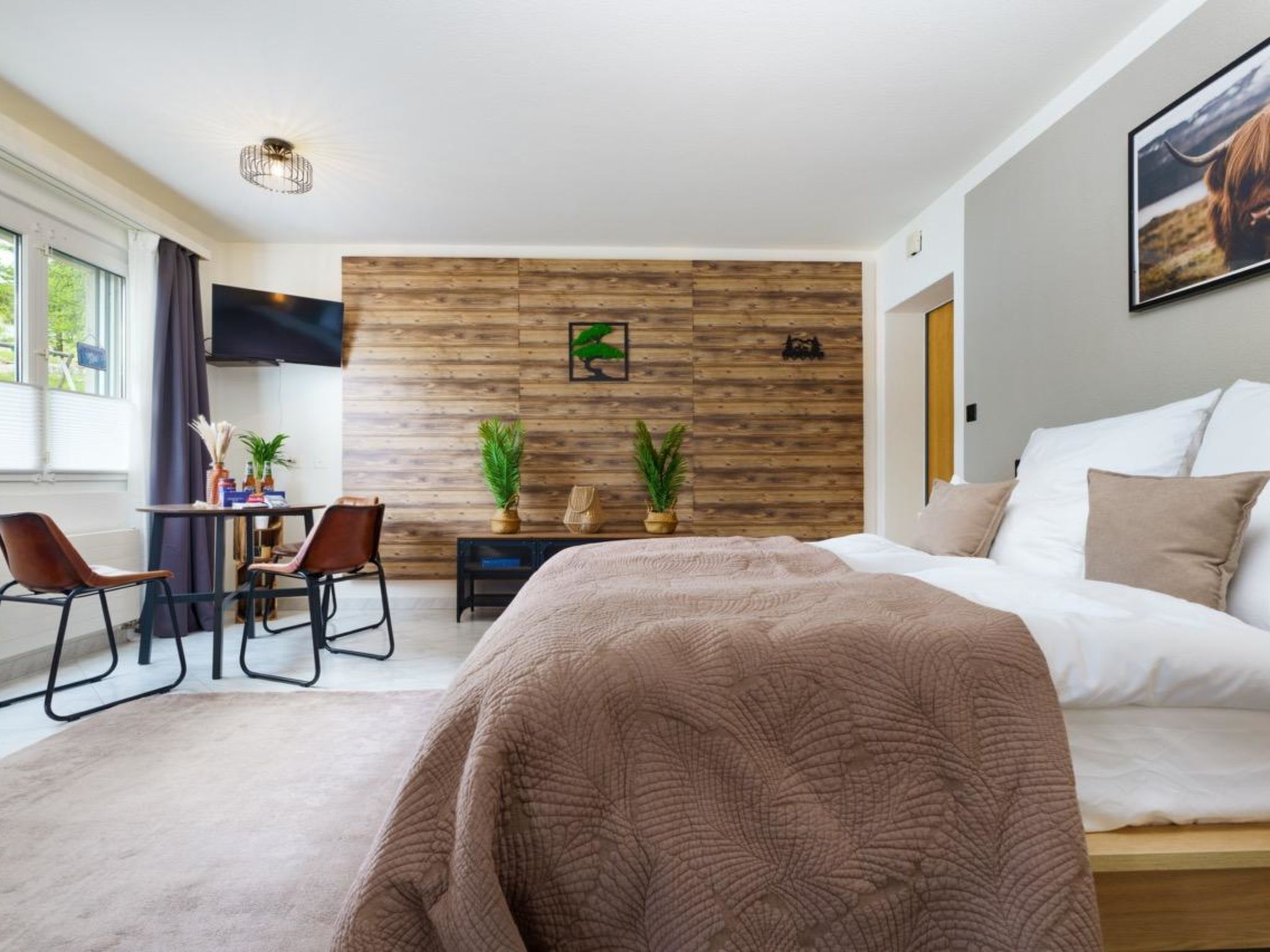 Ein modernes Hotelzimmer mit einem großen Bett, einem Schreibtisch mit zwei Stühlen, einem Fernseher und einer Wandpaneele aus Holz mit einer Zimmerpflanze.