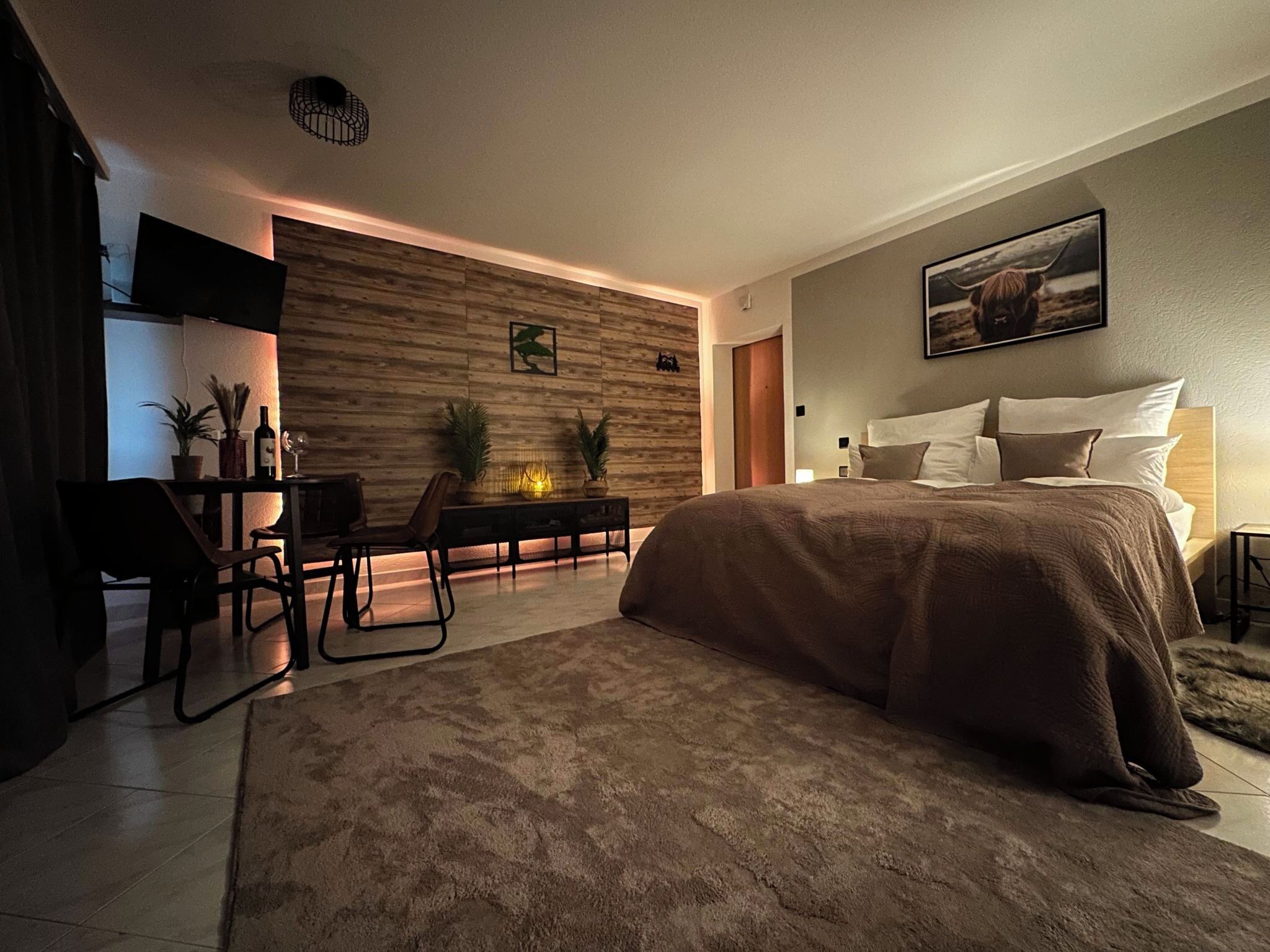 A modern bedroom with a large bed, a desk with a chair, a TV, and a wall-mounted artwork.