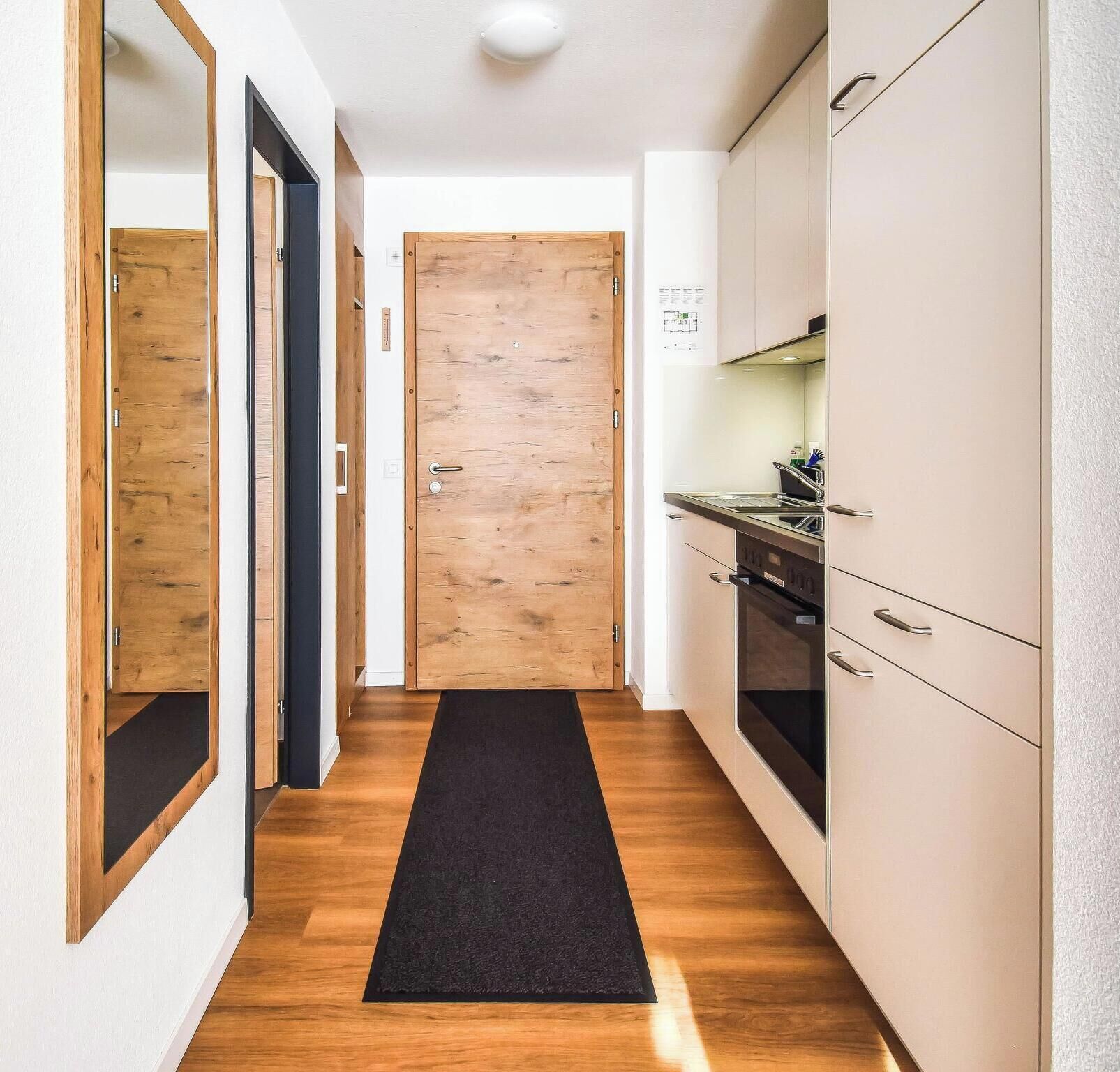 A narrow hallway with wooden flooring, a black mat, and a kitchen area with cabinets and appliances.