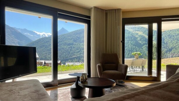 A hotel room with a large window offering a mountain view, a flat-screen TV, a sofa, a chair, and a small table. - Hollywood 1 Appartement/Fewo 6-Bettwohnung by Interhome (Saas-Fee)