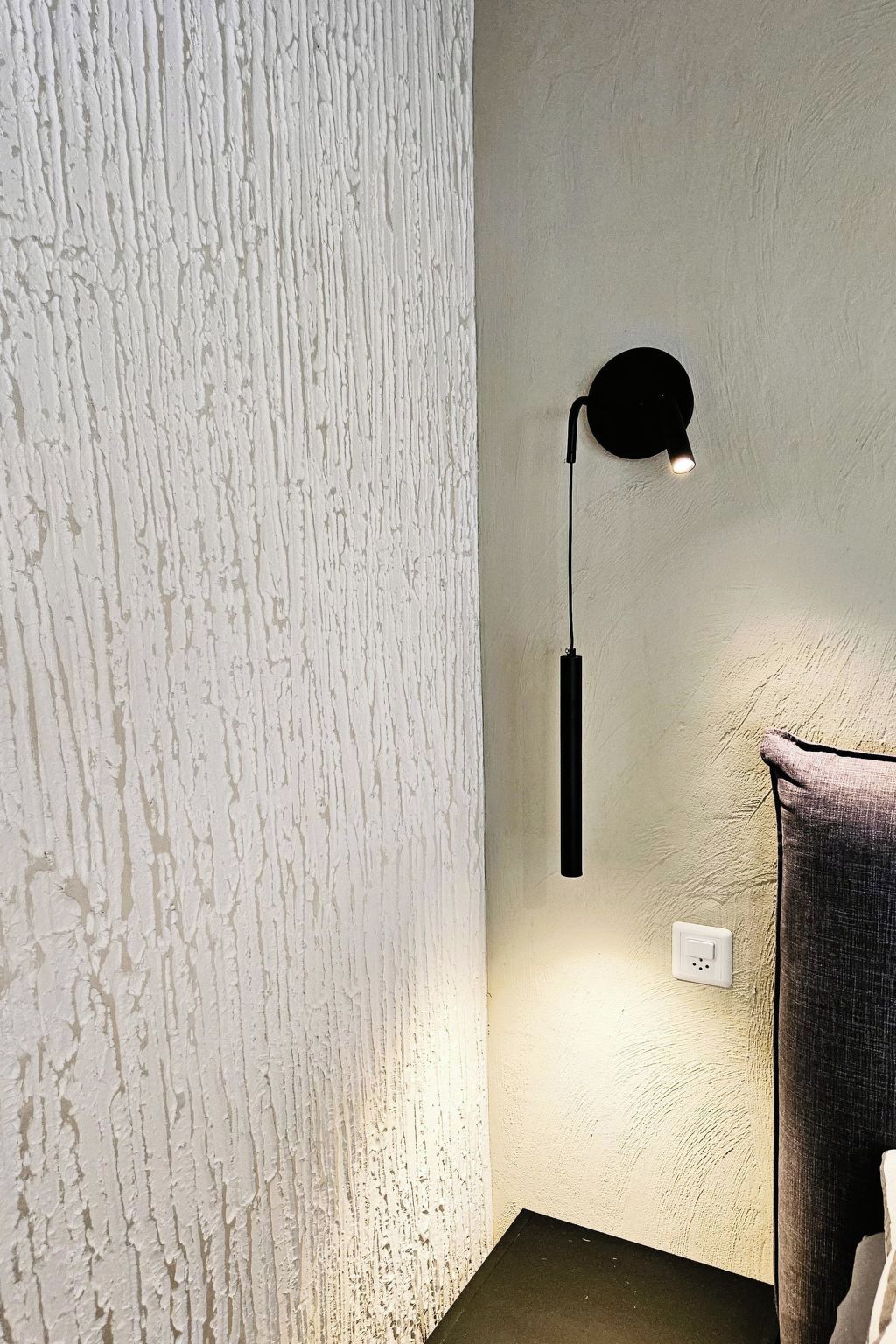A corner of a room with a textured white wall, a black wall-mounted lamp, and a power outlet.