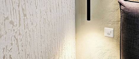 A corner of a room with a textured white wall, a black wall-mounted lamp, and a power outlet.