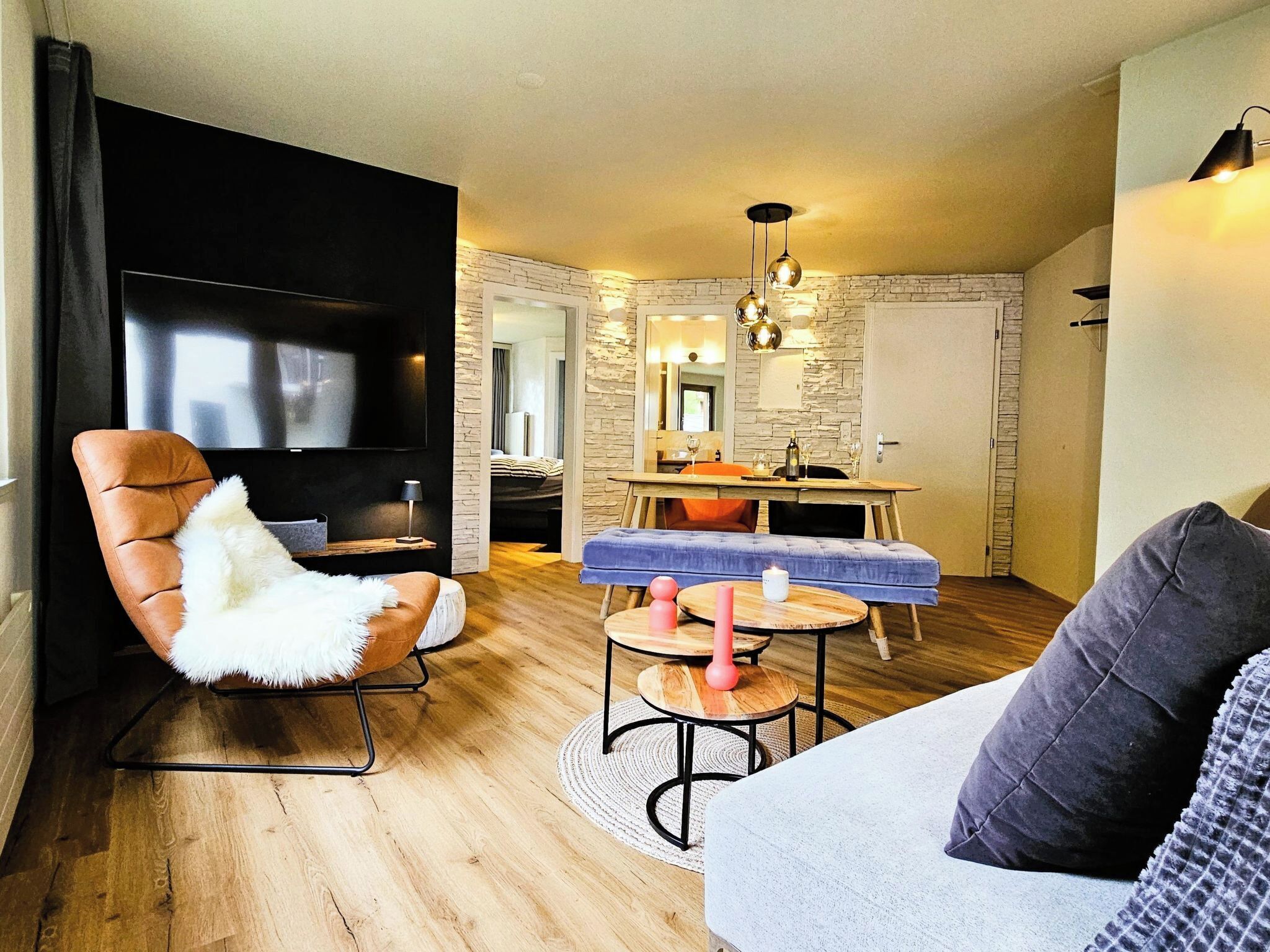 A modern living room with a flat-screen TV, a leather armchair, a sofa, a dining area with a table and chairs, and a stone accent wall.