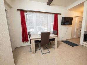 Ein kleiner Essbereich mit Tisch und Stühlen, ein Fenster mit roten Vorhängen und ein an der Wand befestigter Fernseher.