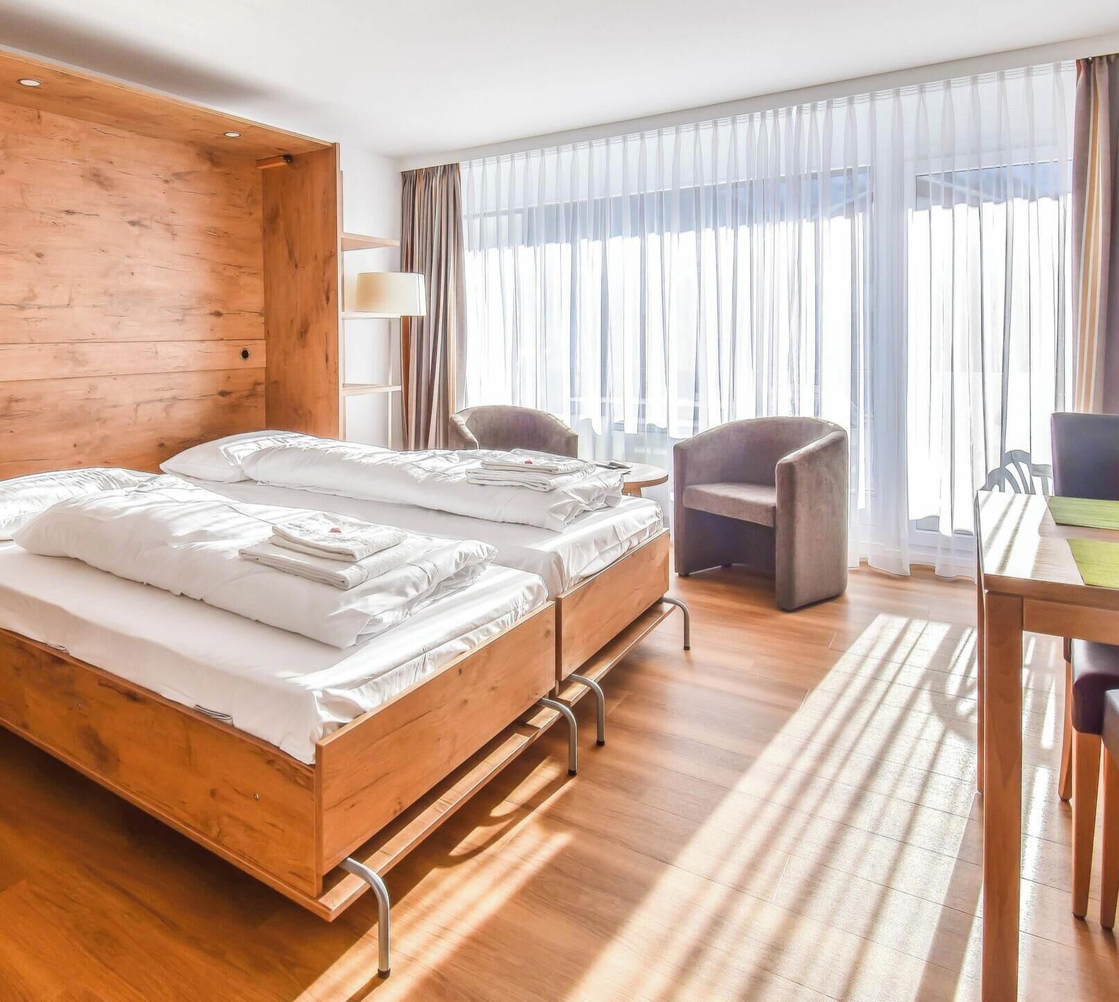A hotel room with two beds, a wooden wardrobe, a desk with a chair, and a large window with curtains.