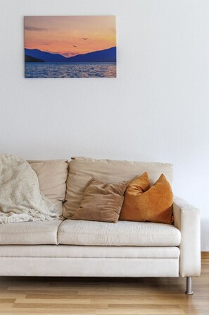 Ein weißes Sofa mit drei Kissen, einer Decke und einem Bilderrahmen mit einem Sonnenuntergang über dem Wasser.