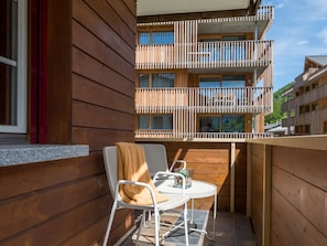 Ein Holzbalkon mit weißem Metallstuhl und Tisch, einem Fenster und Blick auf ein mehrgeschossiges Gebäude.