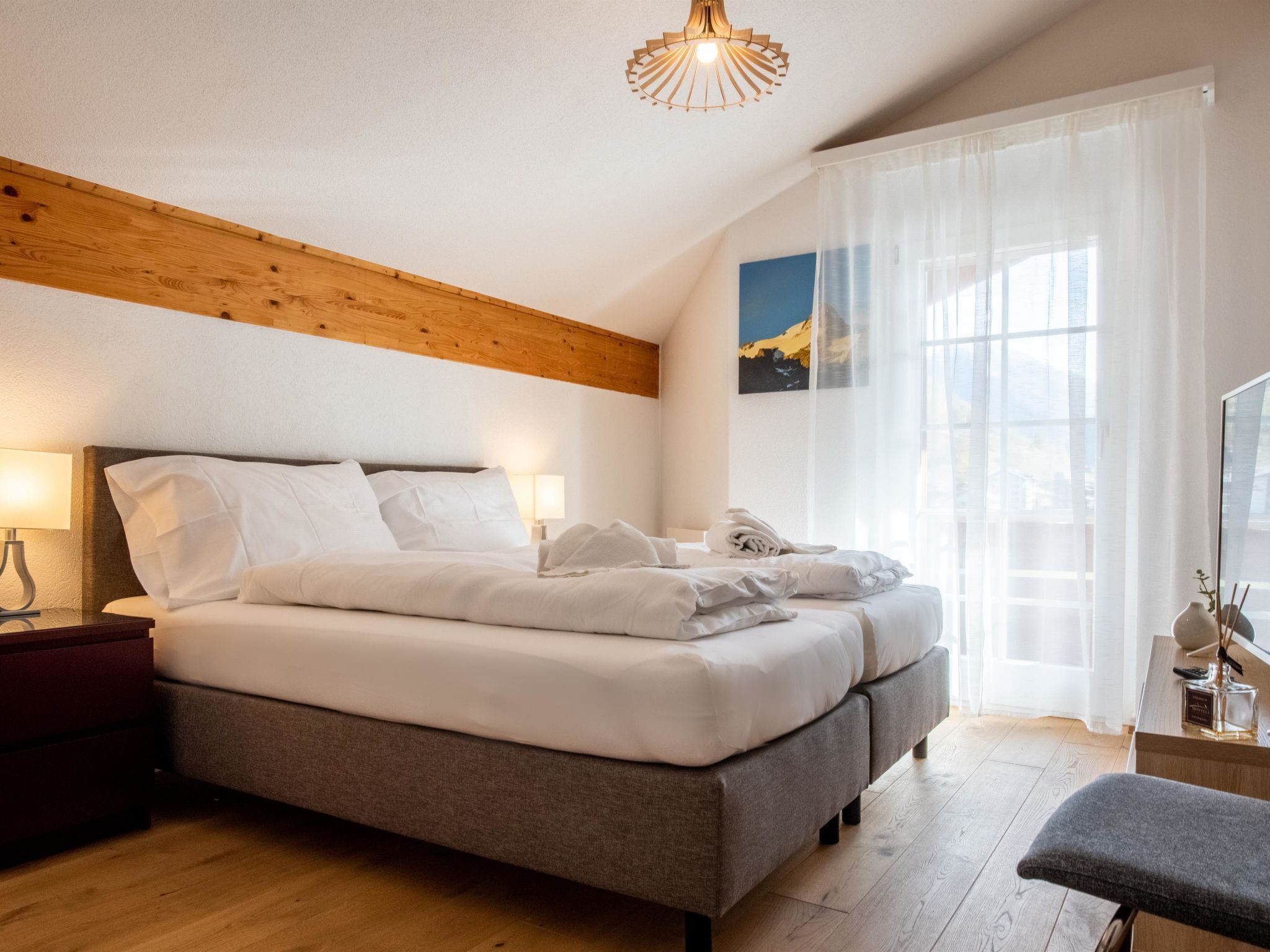 A modern bedroom with a wooden ceiling, a large bed with white linens, a bedside table with a lamp, and a window with sheer curtains.