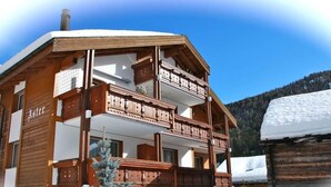Una cabaña de madera con un balcón, un cartel que dice 'Aster' y un paisaje montañoso nevado.