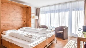 Ein Hotelzimmer mit zwei Betten, einem Holzschrank, einem Schreibtisch mit Stuhl und einem großen Fenster mit Vorhängen.