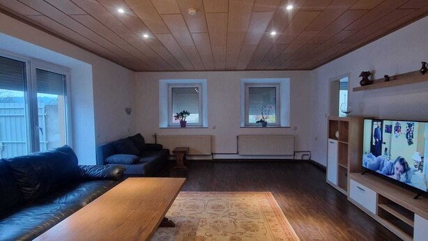 A living room with a wooden ceiling, a black leather sofa, a wooden coffee table, a patterned rug, a television, and a window with blinds.