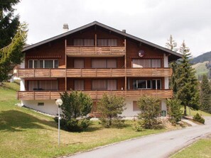 Ein mehrstöckiges Holzgebäude mit Balkonen, umgeben von Grünpflanzen und blauem Himmel.