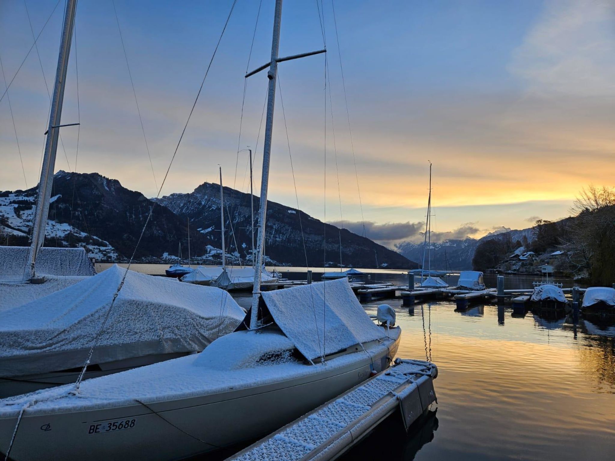Un port de plaisance où les bateaux sont recouverts de neige, l’eau est calme et des montagnes se dressent à l’arrière-plan.