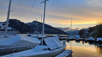 Un port de plaisance où les bateaux sont recouverts de neige, l’eau est calme et des montagnes se dressent à l’arrière-plan.