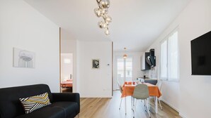 A modern living room with a black sofa, a dining table with orange chairs, and a kitchen area with a black cabinet and a TV mounted on the wall. - Red Maple - Immobiliare Azzurra (Bardolino)