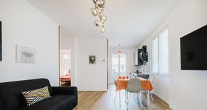 Ein modernes Wohnzimmer mit einem schwarzen Sofa, einem Esstisch mit orangefarbenen Stühlen und einer Küchenzeile mit einem schwarzen Schrank sowie einem an der Wand befestigten Fernseher.
