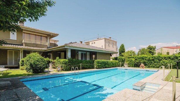 A swimming pool with a clear blue water, surrounded by a well-maintained lawn and a building with a tiled roof. - Red Maple - Immobiliare Azzurra (Bardolino)