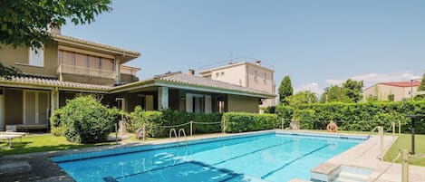 Una piscina con agua cristalina, rodeada por un césped cuidado y un edificio con tejado de azulejos.