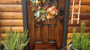 A wooden door with a wreath, a 'FALL' sign, and two potted plants on a wooden deck. - Rustic and Elegant Creekside Log Cabin (Wing)