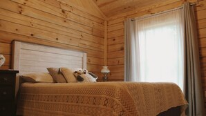 A wooden-paneled room with a bed, a nightstand, and a window with curtains. - Rustic and Elegant Creekside Log Cabin (Wing)