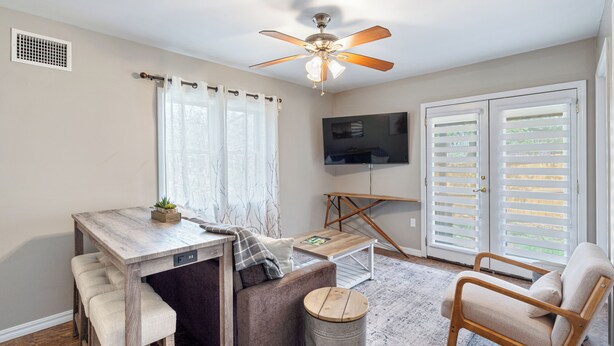 Una habitación con ventilador de techo, televisor, mesa de comedor con bancos, sofá, sillón y ventana con persianas.