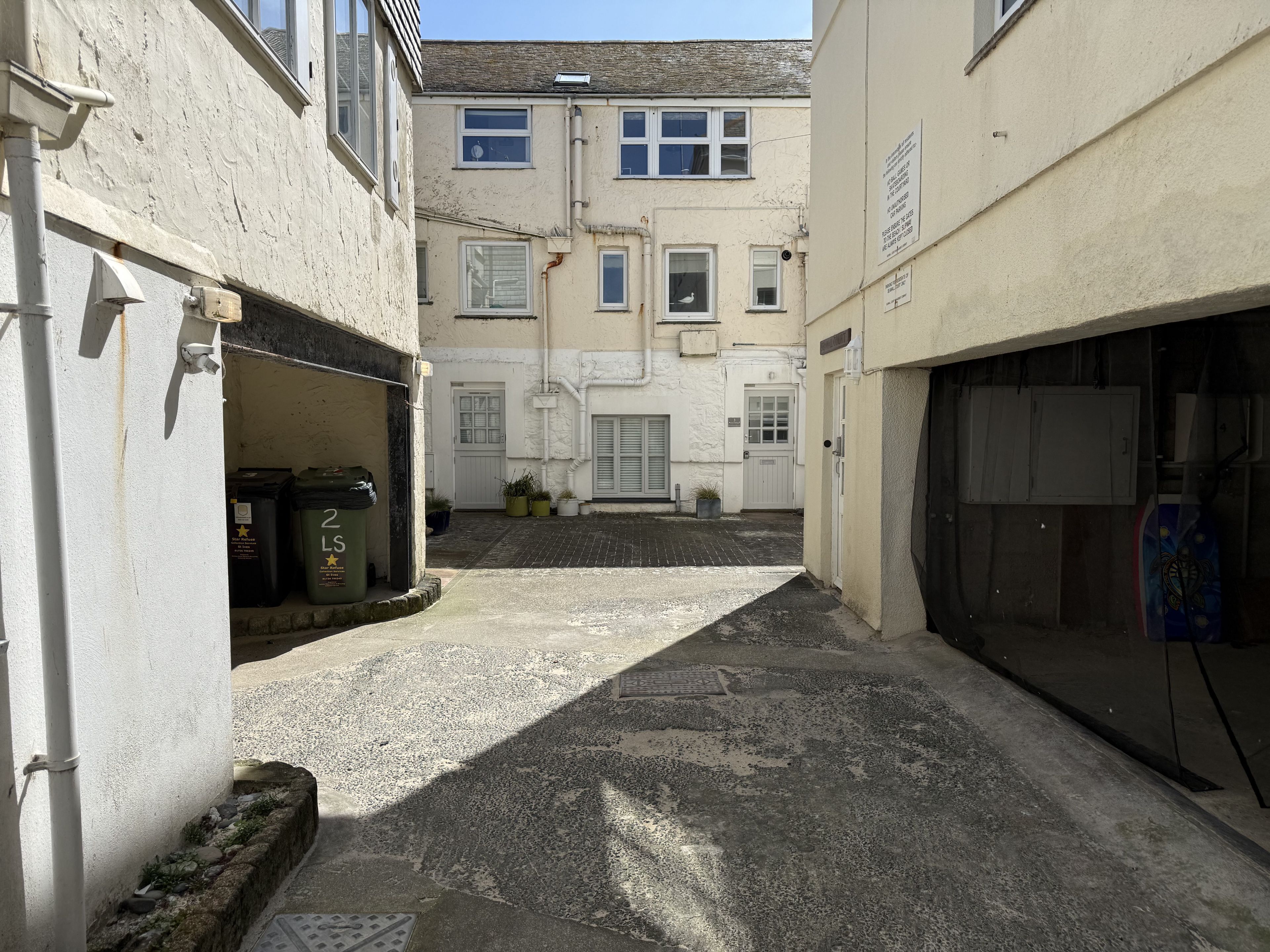 A narrow alleyway between buildings with a cobblestone path, a white door, and a small garden area with potted plants.