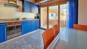 A modern kitchen with blue cabinets, a stainless steel range, and a glass dining table with orange chairs. - Snow-Fun B Penthouse Appartement/Fewo Appartement/Fewo by Interhome (Saas-Fee)