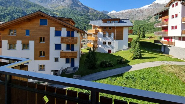 A modern multi-story building with balconies, surrounded by a lush green landscape and mountains in the background. - Snow-Fun B Penthouse Appartement/Fewo Appartement/Fewo by Interhome (Saas-Fee)