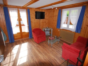 A wooden-paneled room with a TV, red chairs, a glass coffee table, and a window with curtains. - Chalet Alpenruh Appartement/Fewo Nr. 3 Mischabel by Interhome (Saas-Fee)