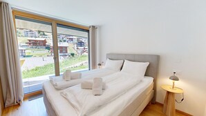 Et hotelværelse med en stor seng, et natbord med en lampe og udsigt til bygninger gennem et stort vindue.