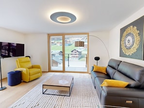 A modern living room with a sofa, armchair, coffee table, and a large window with a view of buildings. - Alphubel Peak & Unlimited Appartement/Fewo Peak 04 by Interhome (Saas-Fee)