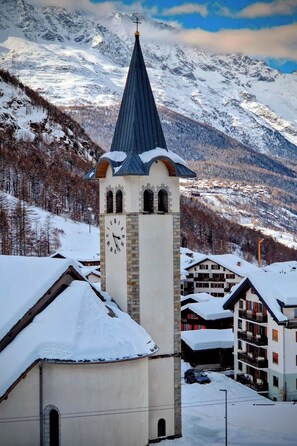 Una església amb una torre del rellotge, envoltada d'edificis i muntanyes coberts de neu.