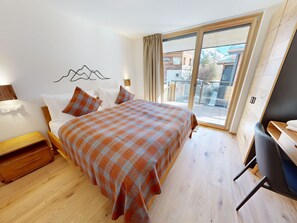 A bedroom with a bed, a desk, a chair, a wooden nightstand, and a window with a view of buildings and mountains. - Alphubel Peak & Unlimited Appartement/Fewo Peak 08 by Interhome (Saas-Fee)